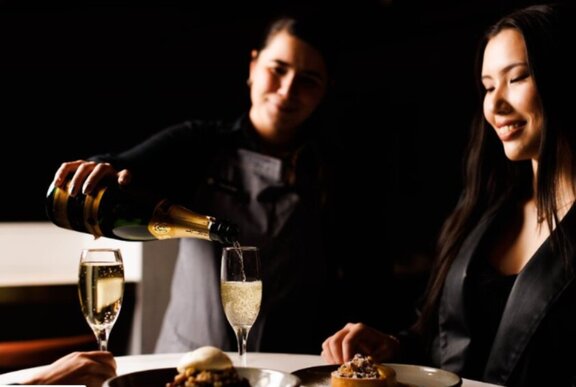 A woman sitting at a table in a restaurant watching a waiter pour her a glass of champagne.