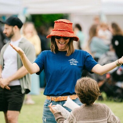 A woman in a blue t-shirt and red bucket hat welcoming a child with arms wide.