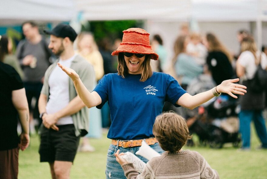 A woman in a blue t-shirt and red bucket hat welcoming a child with arms wide.