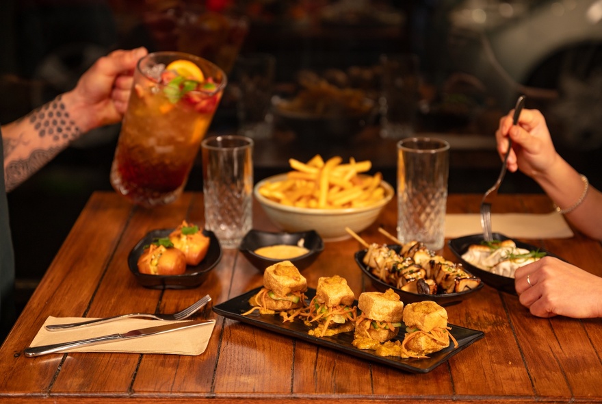 A delicious-looking spread of Asian dishes on a wooden table, with two high-ball glasses and a person's hand pouring a cocktail from a jug.