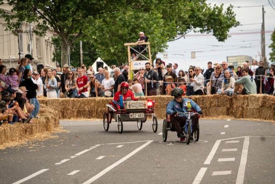 People driving down a closed road in billy carts, with an audience looking on behind hay bales.