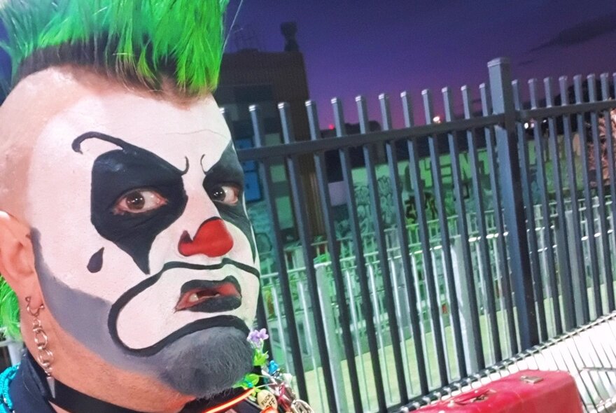An angry looking clown with a green mohawk near a steel fence. 