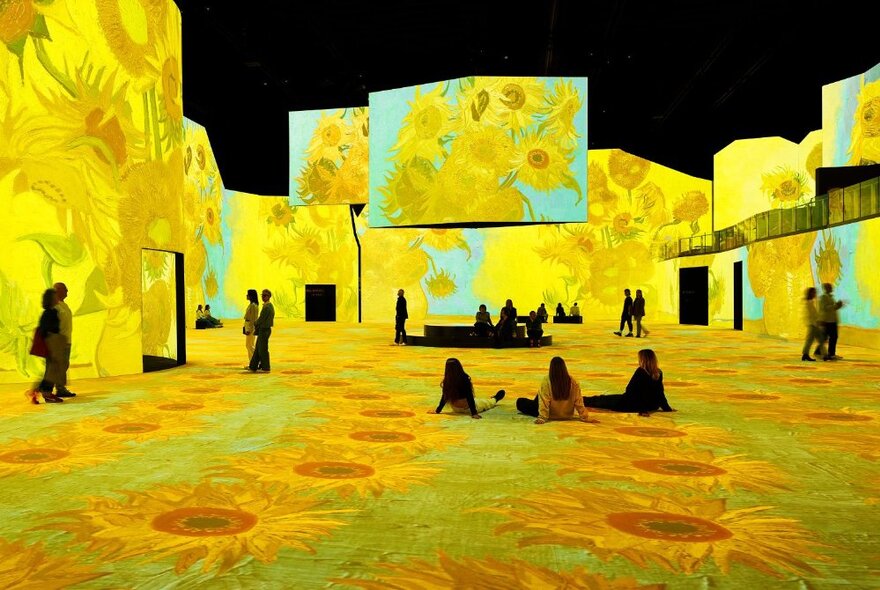 People walking around or sitting on the floor of a very large exhibition space with the Vincent van Gogh's sunflower paintings digitally projected on all the wall and floor surfaces to create an immersive experience.