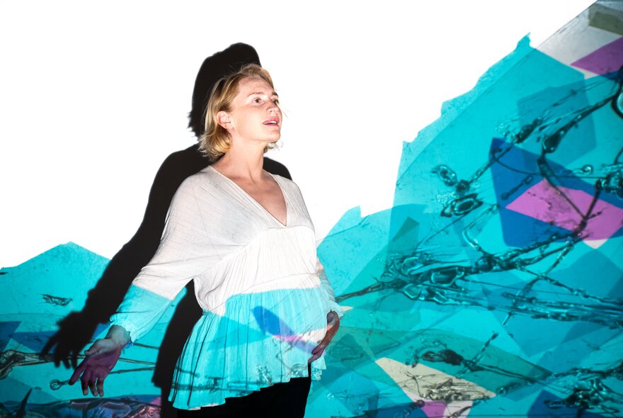 A person standing with one hand on their pregnant belly, illuminated by projected blue and pink toned shapes. 