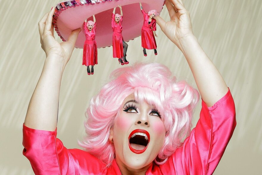Comedian wearing pink blonde wig, holding a hat with tiny figures of herself above her head.