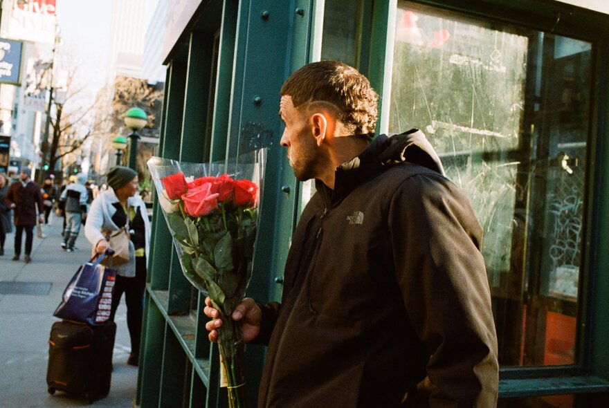A man standing on a New York city sidewalk holding a large bouquet of red roses near a green subway kiosk.