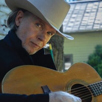 Australian singer-songwriter Rick Price, wearing a ten-gallon cowboy hat and strumming a guitar, seated outside.