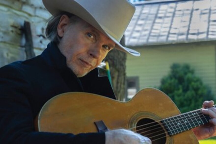Australian singer-songwriter Rick Price, wearing a ten-gallon cowboy hat and strumming a guitar, seated outside.