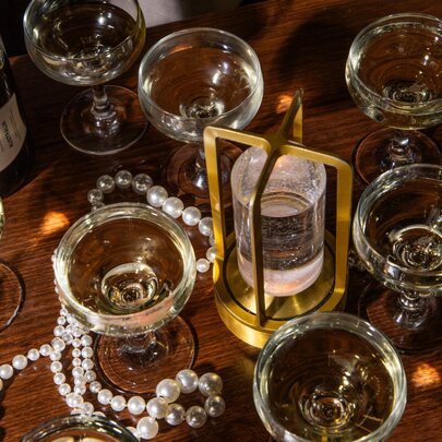 Several coupe glasses of sparkling wine, a gold-toned LED-lit stand, and pearl decorations on a wooden table.