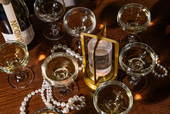 Several coupe glasses of sparkling wine, a gold-toned LED-lit stand, and pearl decorations on a wooden table.