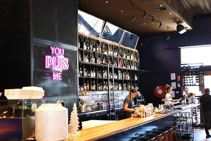 Bar counter with staff preparing drinks, shelves of bottles and pink neon signage.