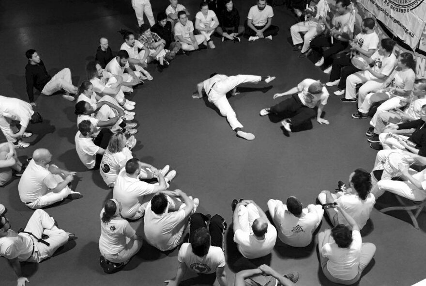View from above of people participating in a Capoeira Angola class, with two people dancing and moving in the centre of a seated group of onlookers. 