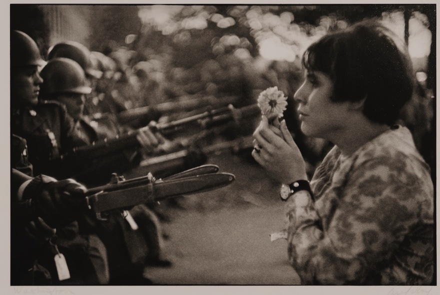 Woman holding a flower in front of a row of soldiers holding pointed guns with bayonets.