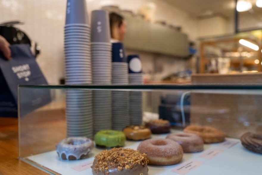 A glass countertop display case with an assortment of different donuts inside; takeaway coffee cups stacked behind it on the counter.