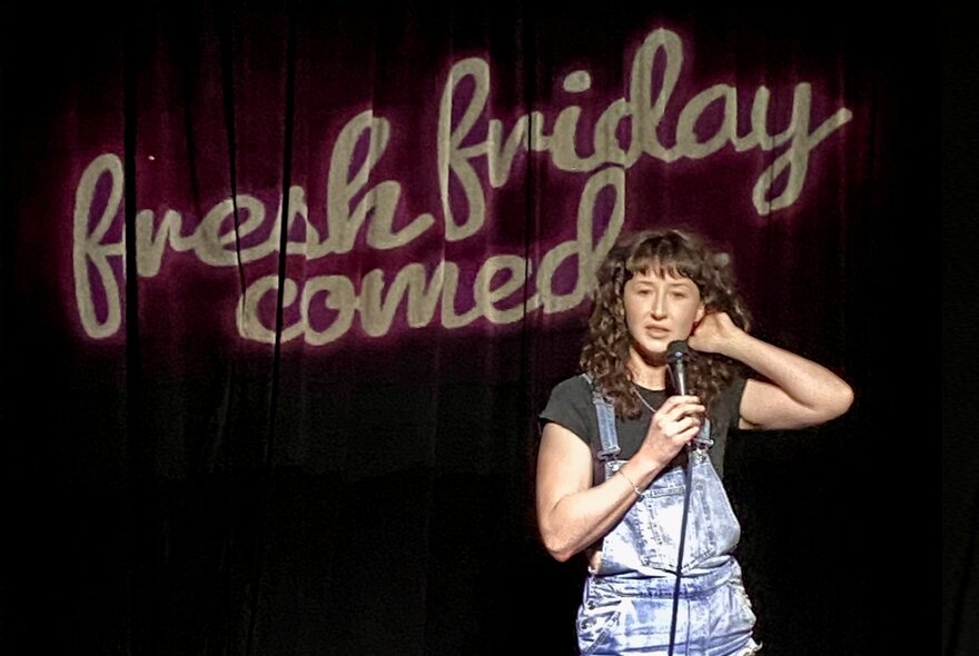 A comedian performing stand-up on a stage, holding a microphone, the black curtain behind them displaying the words 'fresh friday comedy' in cursive font.