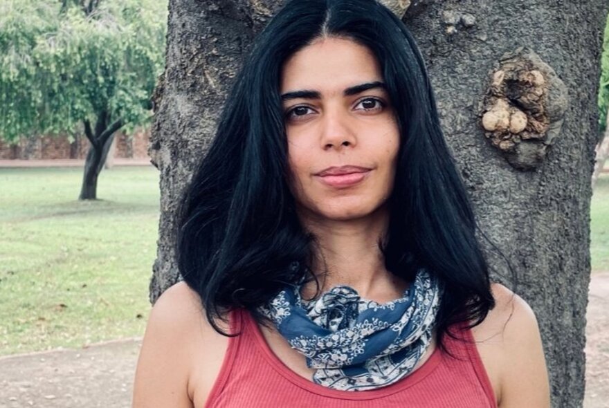 Graphic novelist Ita Mehrotra, a woman standing outside in front of a tree wearing a pink sleeveless top, a blue cotton scarf around her neck and her straight black her framing her face.