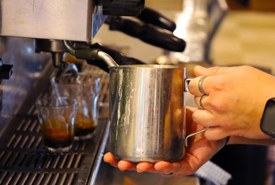 A hand holding a small stainless steel milk jug and heating it up using the steamer arm on a coffee machine.  