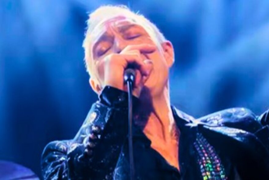 A man with short blond hair and his eyes closed singing into a microphone on a blue backlit stage.