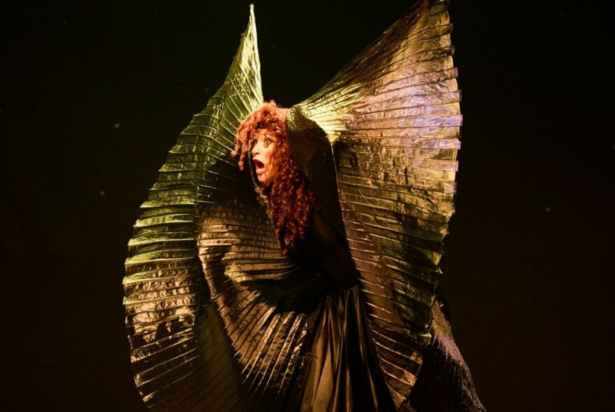 UK performer, Sarah-Louise Young, dressed in a bronze coloured ruffled costume, performing in character as Kate Bush on a darkened theatre stage.