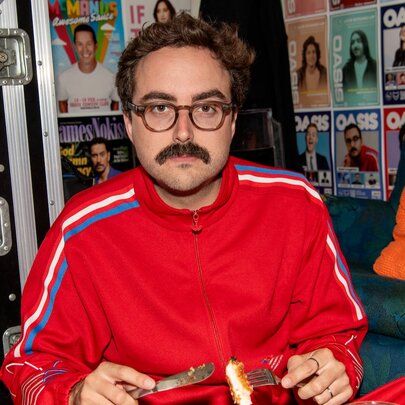 Stand up comedian, Daniel Muggleton, wearing a red tracksuit and glasses while holding a knife and a fork with a piece of crumbed chicken on it, in a pub setting. 