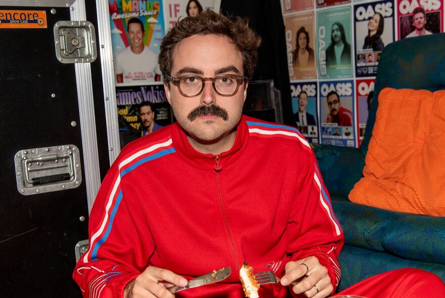 Stand up comedian, Daniel Muggleton, wearing a red tracksuit and glasses while holding a knife and a fork with a piece of crumbed chicken on it, in a pub setting. 