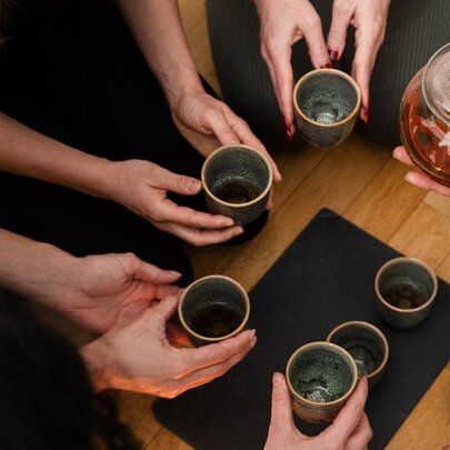 People gathered in a circle holding cups in front of a glass teapot.