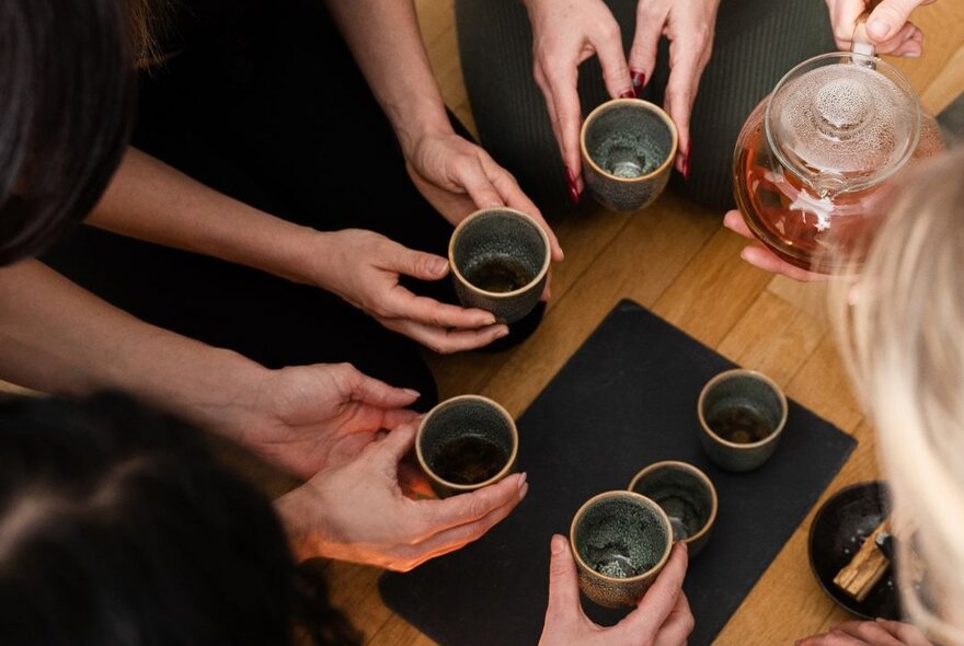 People gathered in a circle holding cups in front of a glass teapot.