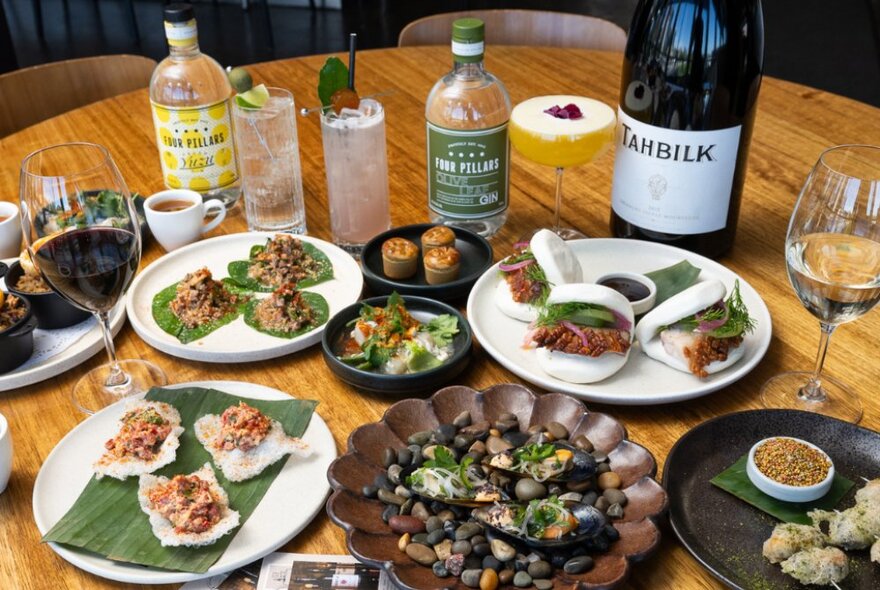 A selection of carefully arranged food on plates,  glasses and bottles of wine and gin behind the plates, on a round wooden table in a restaurant.