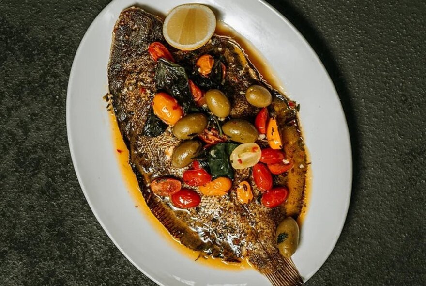 A pan-fried whole fish presented on an oval white plate with an olive and cherry tomato sauce.