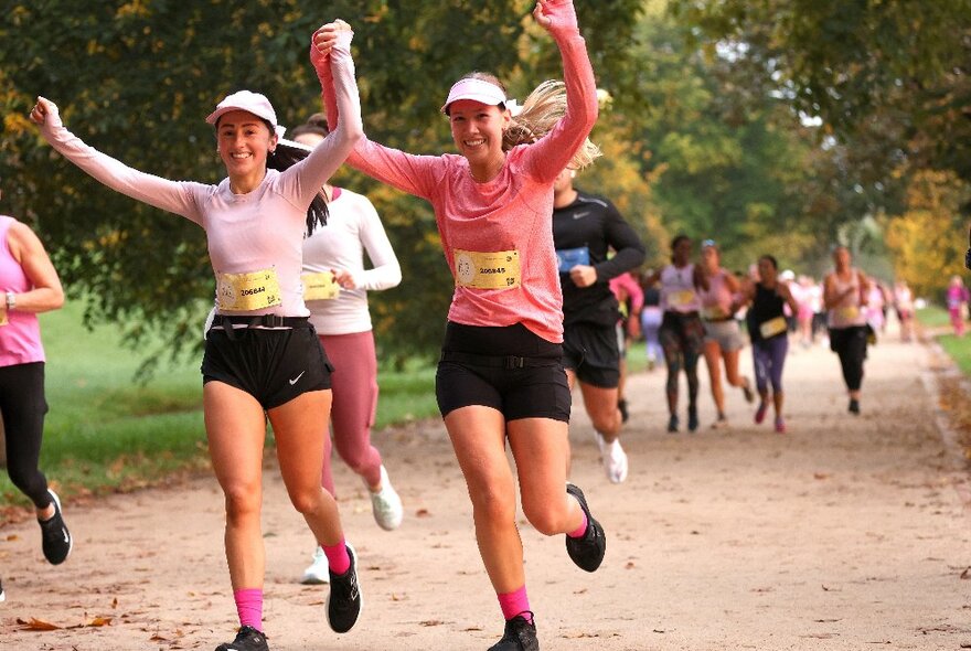 Two runners participating in the Mother's Day Classic fun run, wearing pink tops and holding up their arms in a cheering gesture as they jog on a path.