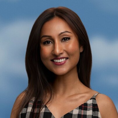 Comedian and writer Urvi Majumdar, smiling at camera, with long straight dark brown hair, she wears a check dress with thin shoulder straps and stands in front of a blue sky background.