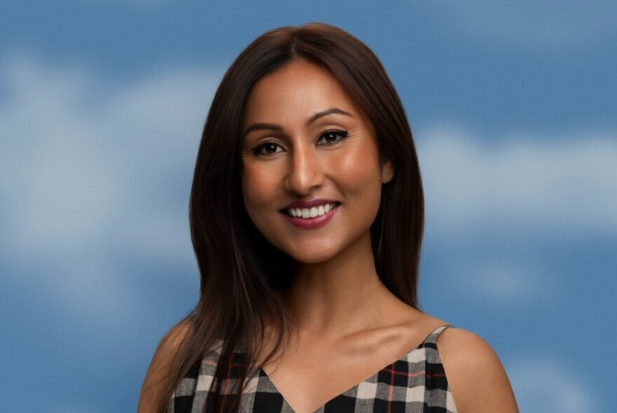 Comedian and writer Urvi Majumdar, smiling at camera, with long straight dark brown hair, she wears a check dress with thin shoulder straps and stands in front of a blue sky background.