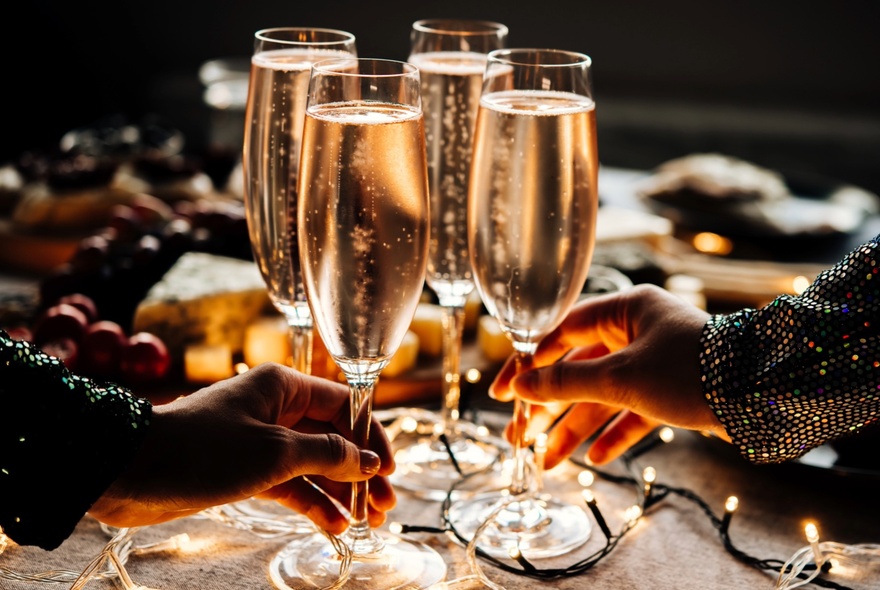 Two hands selecting two flute glasses of sparkling wine from a table decorated with fairy lights.