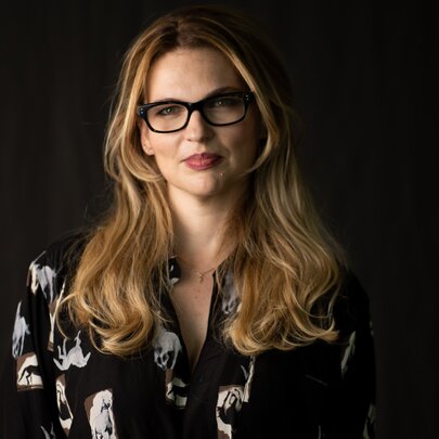 Journalist Amy Remeikis wearing glasses and sitting with a neutral expression in a black room. 