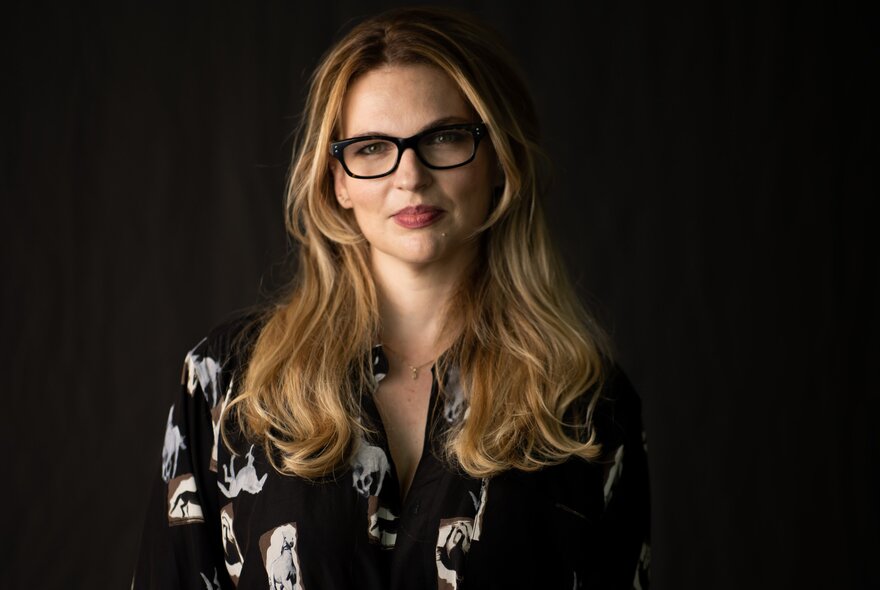 Journalist Amy Remeikis wearing glasses and sitting with a neutral expression in a black room. 