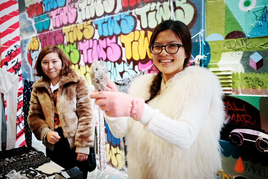 Two women at a secondhand staff with posters and graffiti on the wall behind them.
