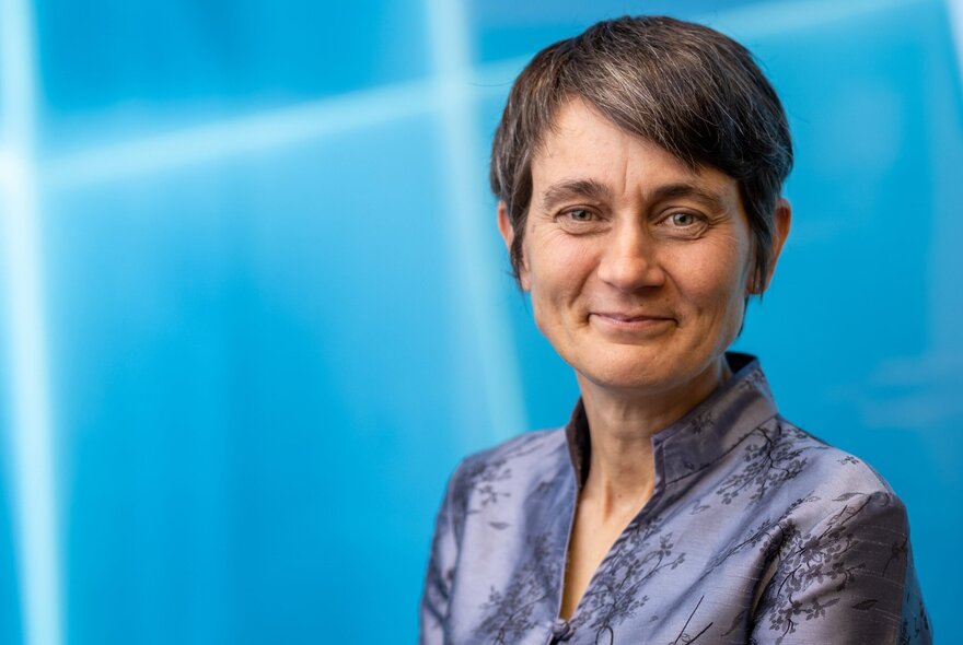 Older woman with short hair and blue blouse, against blue background.