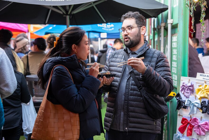 Two people chatting while eating at an outdoor market, a large black umbrella behind them. 