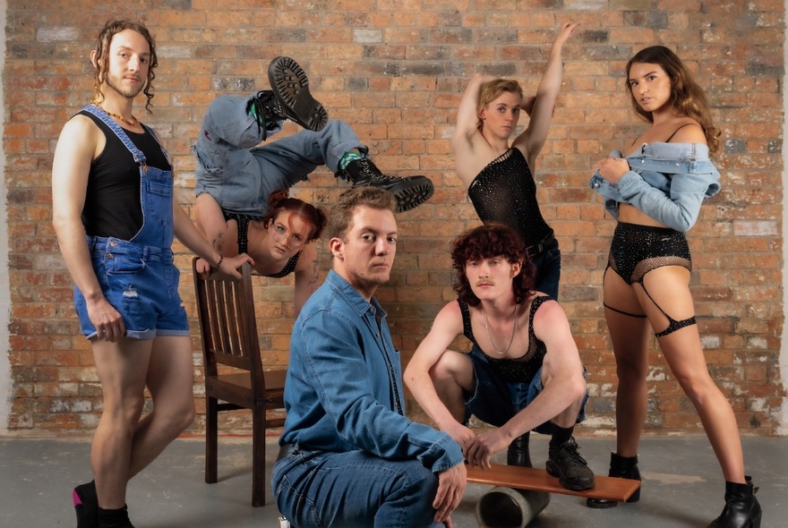 A cast of circus performers looking at the viewer and all striking a different pose, in a room with a brick wall behind them.