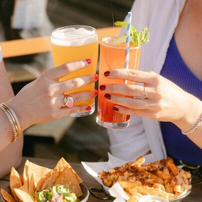 Hands clinking orange drinks over bowls of food in a sunny setting.