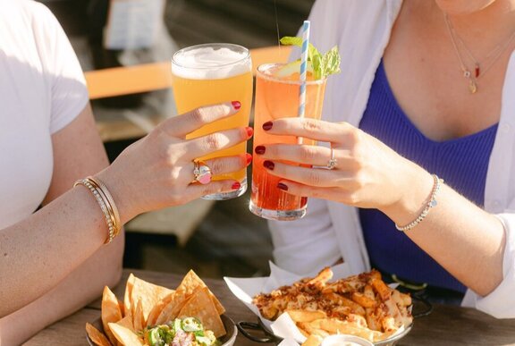Hands clinking orange drinks over bowls of food in a sunny setting.