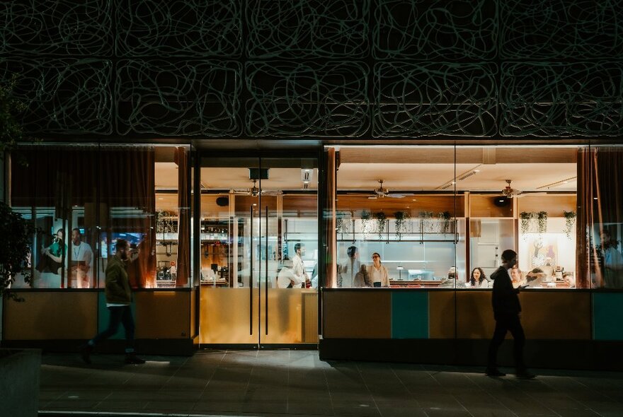 The exterior of Barra restaurant, showing a pedestrian walking past a lit up window with diners sitting inside.