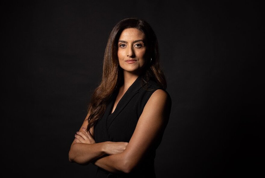 Journalist Nour Haydar, her arms crossed in front of her, looking directly at the viewer and standing in front of a black background.