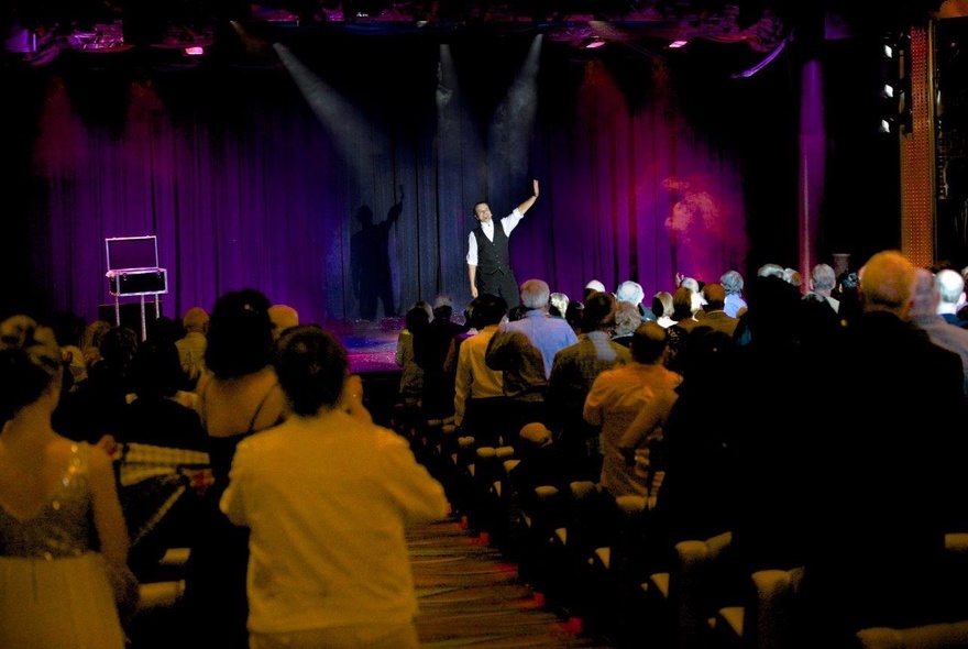 Audience seated in a venue watching a magician wearing a waistcoat performing on stage.