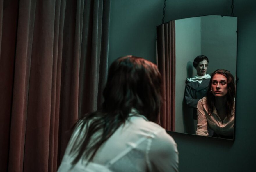 A woman seated in front of a mirror and curtains, another woman visible in the mirror.