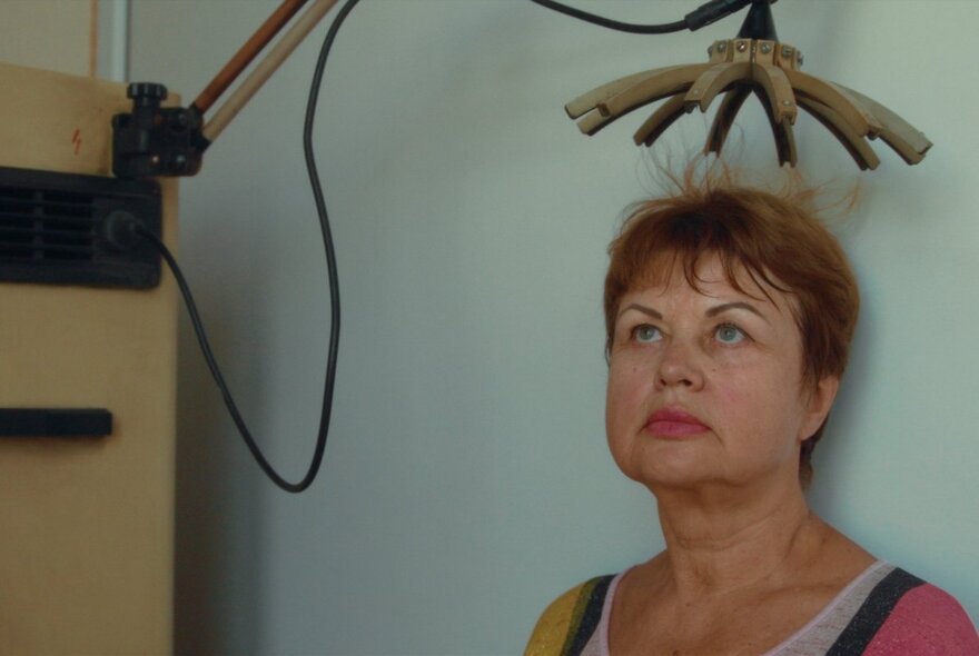 A middle-aged woman with her back against a pale blue wall, a static electricity device above her head making her hair stand on end.