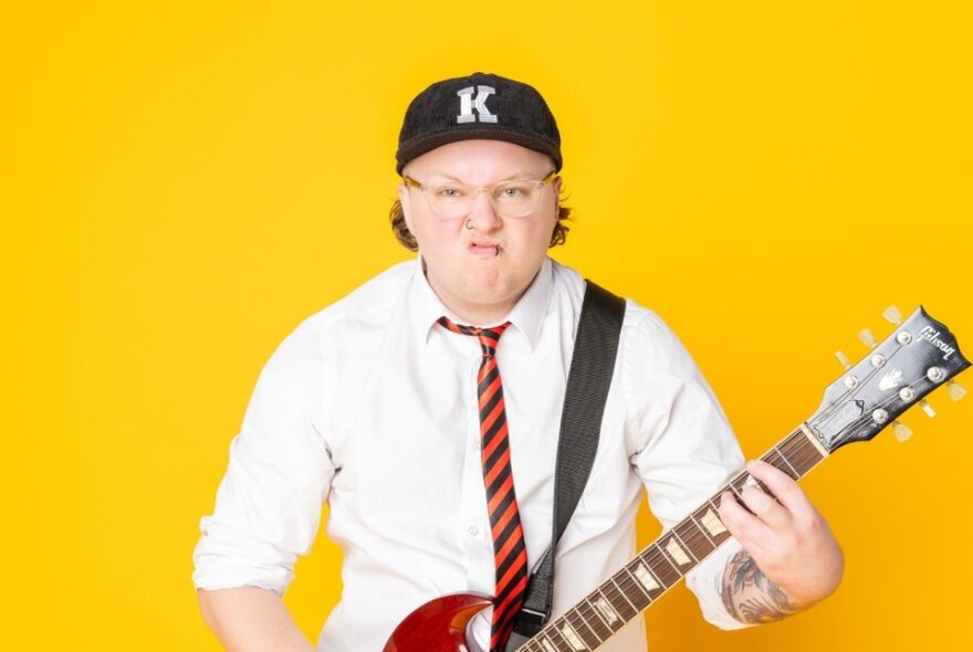 Comedian, Kaileb Hyland, wearing a white shirt, school tie, baseball cap and glasses, playing a guitar in front of a yellow background.