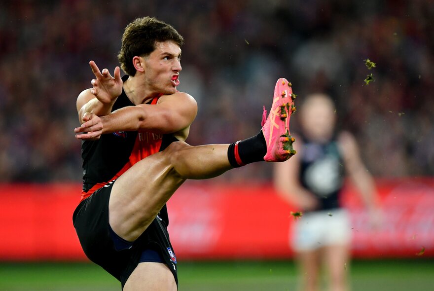 An Essendon player moments are a kick with his leg still high in the air and pink boots on. 
