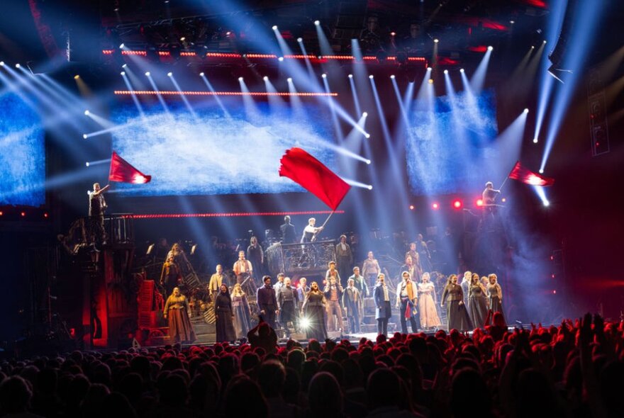 Large ensemble cast singing and performing on stage in the musical Les Misérables The Arena Spectacular, with stage spotlights and smoke haze.