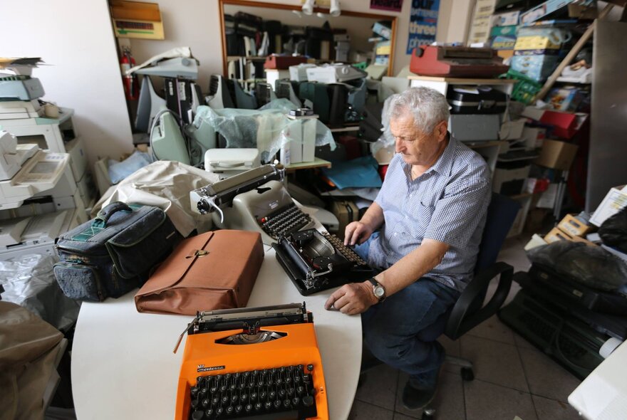 A man in a room crowded with things, sitting at a table with three typewriters on it.